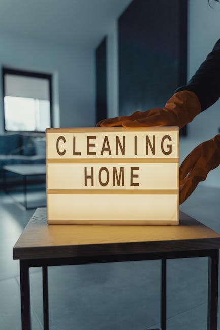 A person wearing yellow cleaning gloves holding a illuminated sign with black letters spelling 'CLEANING HOME' on a wooden surface. The background features a blurred interior space with natural light